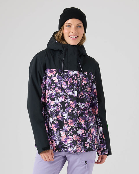 A person wearing a black beanie and the Roxy Jetty Jacket in Blended Bouquet—a women’s snowboard jacket with a floral pattern in shades of purple, pink, and black—stands against a plain light background, smiling at the camera. The Jetty Jacket features warm insulation and is made from recycled materials for comfort and sustainability.