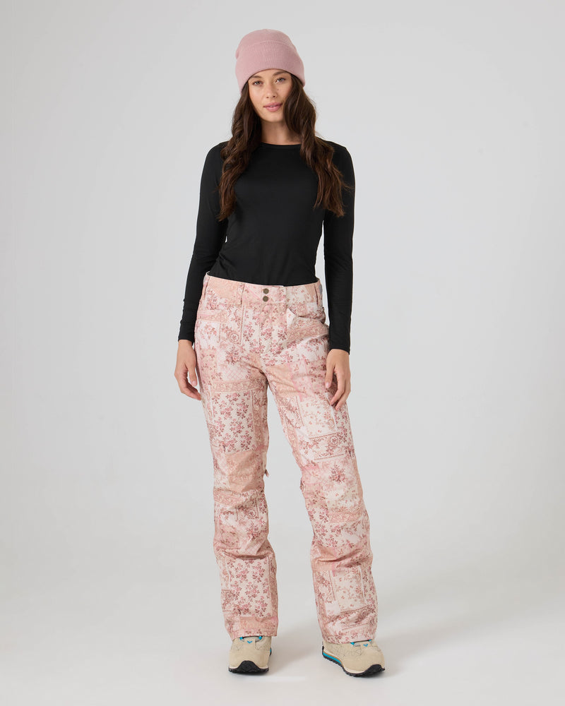 A person wearing a pink beanie, black long-sleeve shirt, and Roxy Backyard Pants in Earthy Wahine—a pair of women’s snowboard pants crafted from recycled fabric with waterproof protection—stands facing forward against a plain light background, finished with beige shoes.