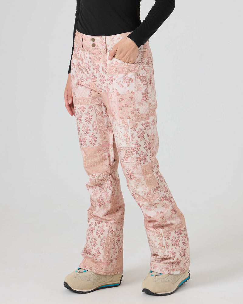 Wearing the Roxy Backyard Pants—pink floral patterned women’s snowboard pants crafted from recycled fabric—paired with a black long-sleeve shirt and beige shoes, a person stands with one hand in their pocket against a plain white background, ready for waterproof protection in style.