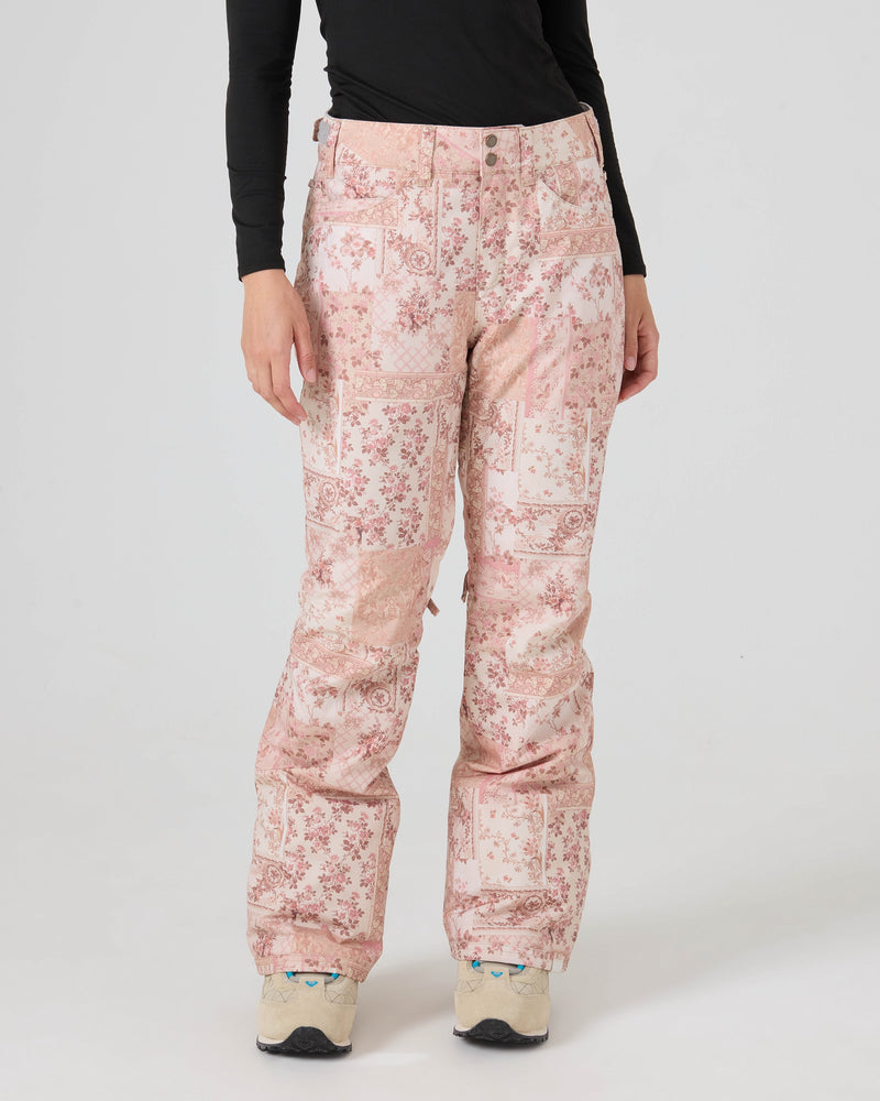 A person wearing Roxy Backyard Pants in a light pink pattern made from recycled fabric, paired with a black long-sleeve shirt and white sneakers, stands against a plain light gray background. The person’s head is not visible in the image.