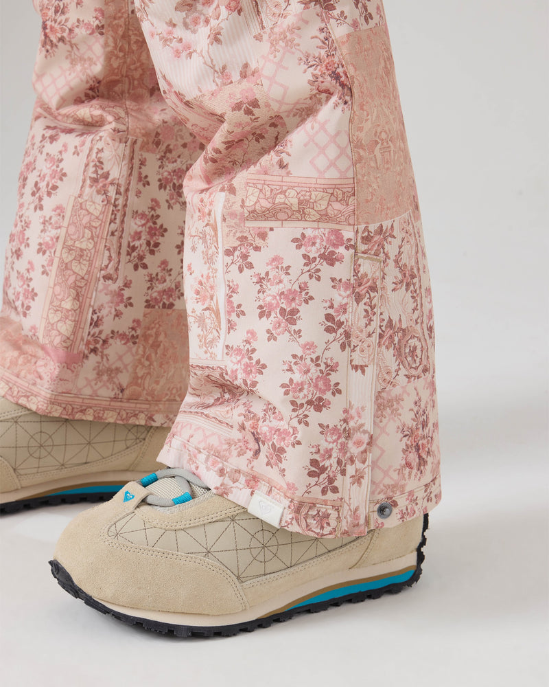 A close-up of a person wearing beige sneakers with blue accents and Roxy’s Backyard Pants—pink floral-patterned pants made from recycled fabric—standing on a white background.