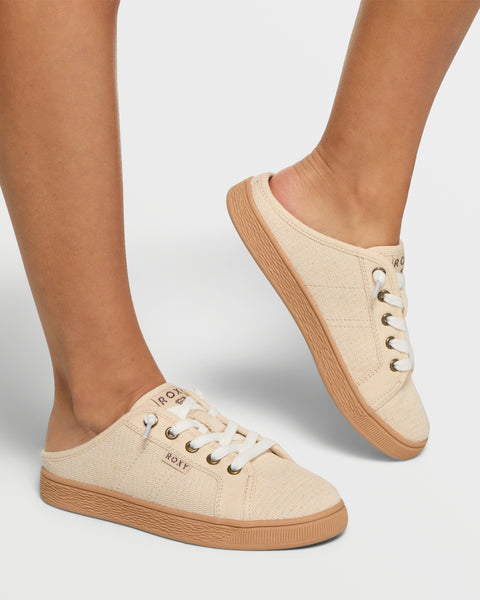 A person wearing Roxy Sunny Stroll Mule Sneakers in beige with white laces, light brown soles, and ROXY branding on the side and tongue, standing on a white background.
