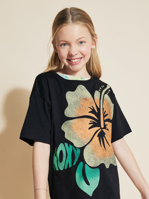 A smiling girl with long blonde hair wears the Roxy Girls' Hibiscus Oversized Tee, a black shirt with a vibrant tropical graphic and green brand name, standing against a plain beige background.