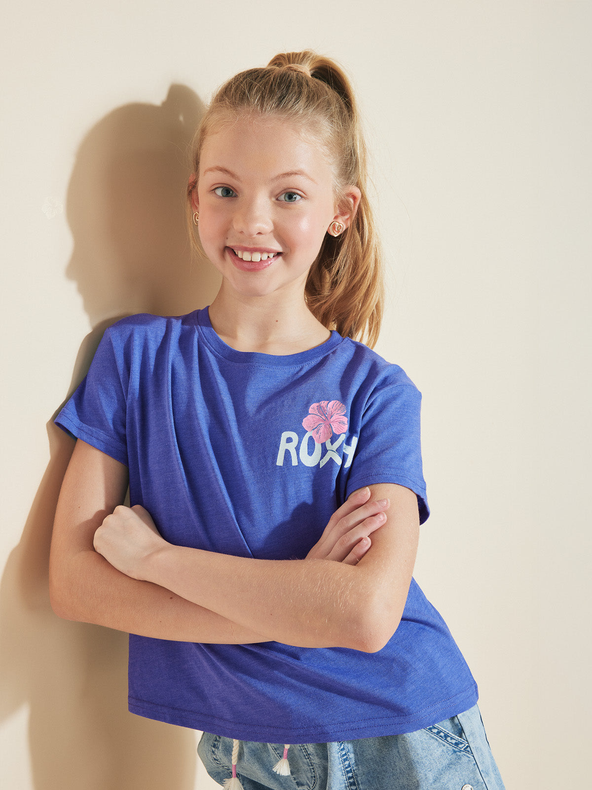 A smiling girl with blonde hair in a ponytail wears the ROXY Girls' Surf Trip Tee—a soft cotton jersey with a pink flower graphic—styled oversized with light denim shorts. She stands against a light wall, arms crossed.