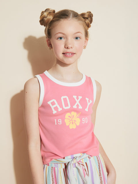 A young blonde girl with double buns wears the Roxy Girls' Collegiate Arch Tank, featuring “ROXY 1990” and a yellow flower, paired with striped pants. She smiles softly in front of a neutral background.