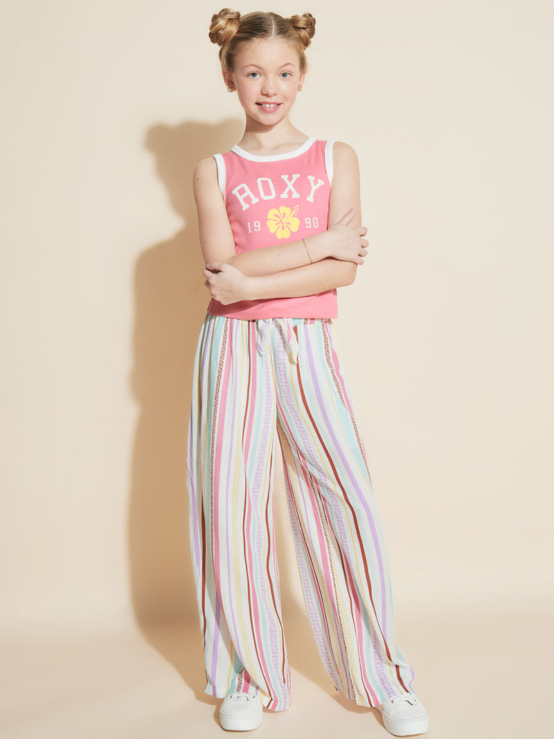 A smiling girl with blonde hair in two buns wears a pink tank top, white sneakers, and Roxy Girls' Bali Stripe Pant in pastel stripes, standing against a beige background with her arms crossed.