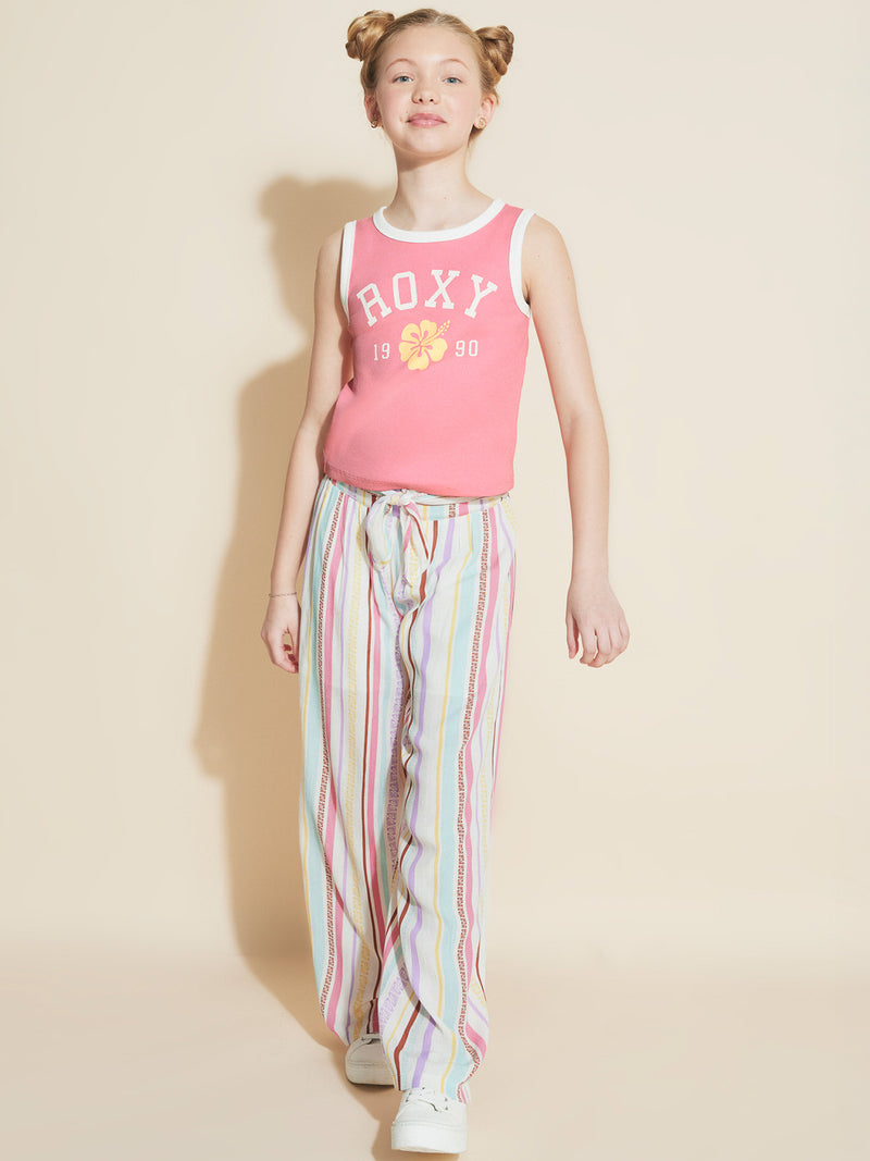 A smiling young girl with blonde hair in two buns walks forward against a beige background, wearing a pink tank top and Roxy Girls' Bali Stripe Pant.