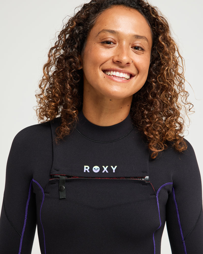 A smiling person with curly hair wears the 3/2mm Elite Xtra Stretch Front Zip Wetsuit by Roxy. The background is plain and light-colored.
