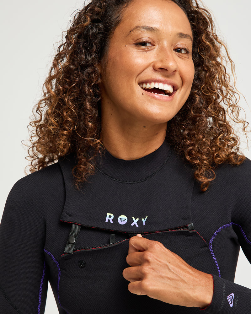A person with curly hair smiles while adjusting the front zipper of the 3/2mm Elite Xtra Stretch Front Zip Wetsuit. The wetsuit is black with Roxy branding, set against a plain, light background.