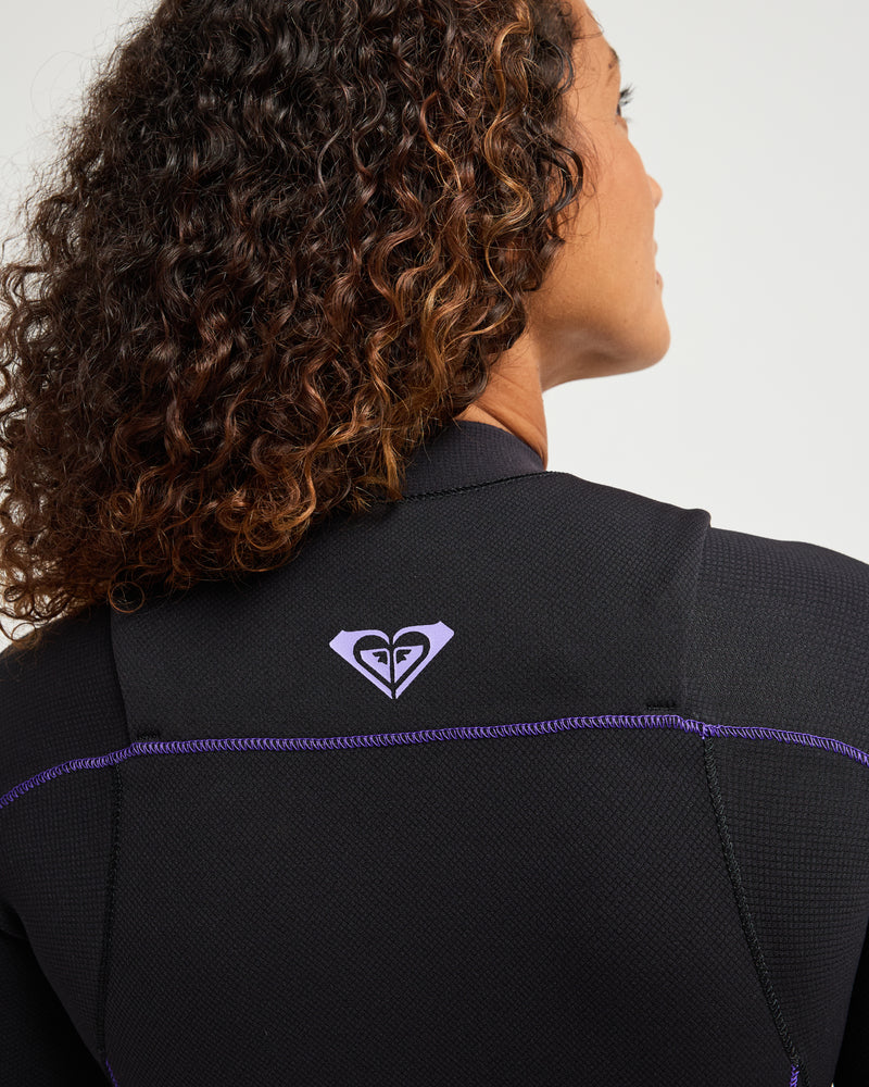 A person with curly hair wears the Roxy 3/2mm Elite Xtra Stretch Front Zip Wetsuit, which is black with purple stitching and a purple heart-shaped logo on the back.