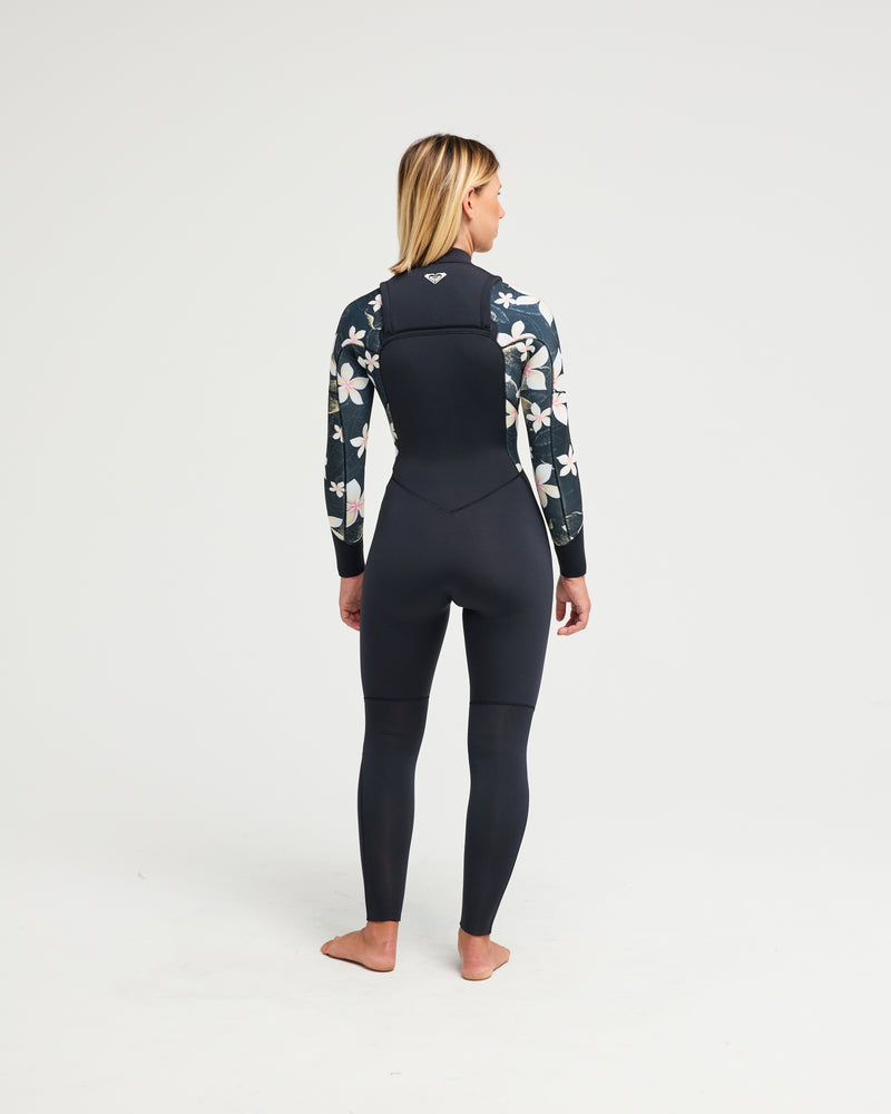 A woman stands barefoot, facing away in the Roxy 4/3mm Swell Natural Front Zip Wetsuit, featuring black fabric with floral patterns on the sleeves and shoulders against a plain white background.
