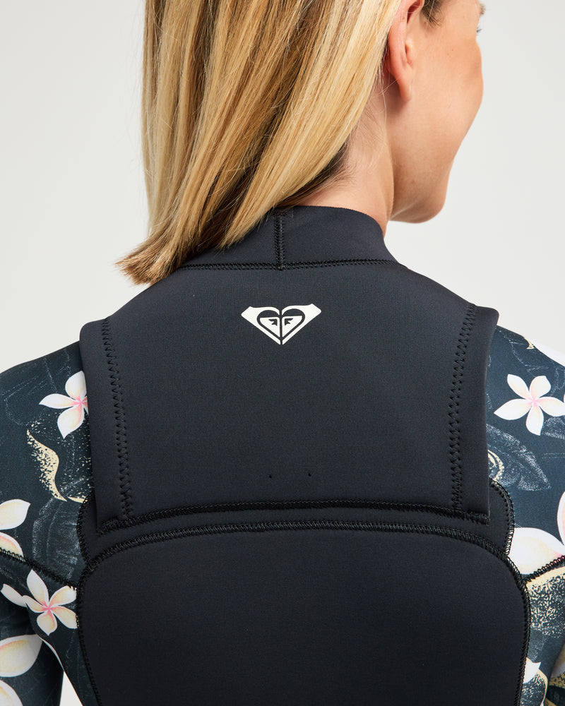 A woman with blonde hair wears the 4/3mm Swell Natural Front Zip Wetsuit by Roxy, featuring a black design with a white Roxy logo on the back and floral-patterned sleeves.