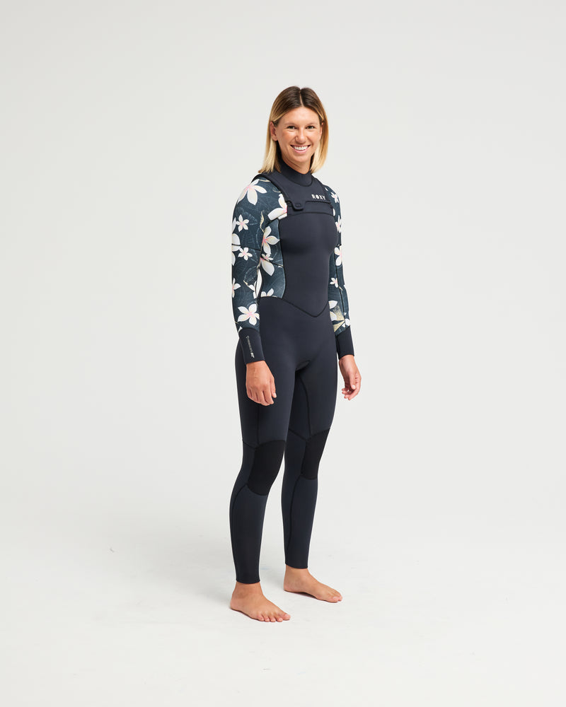 A woman stands barefoot on a white background, smiling and relaxed, wearing the Roxy 4/3mm Swell Natural Front Zip Wetsuit with black fabric and floral-patterned sleeves and shoulders.