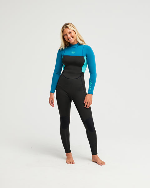 A woman stands barefoot, smiling, and wearing the Roxy 3/2mm Prologue Back Zip (Flatlock) Wetsuit in Baltic against a plain white background.