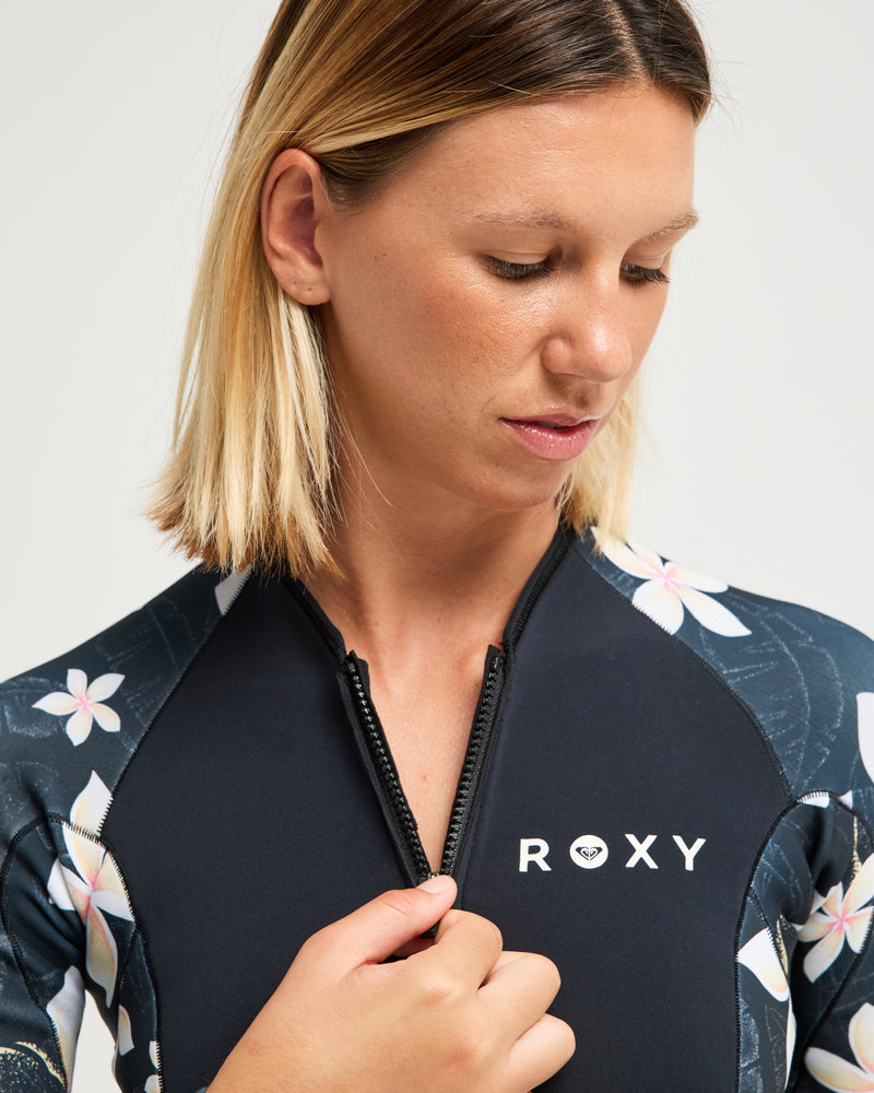 A woman with straight, shoulder-length blonde hair looks down while unzipping a black floral 2mm Swell Natural Back Zip Spring Suit Wetsuit by Roxy.