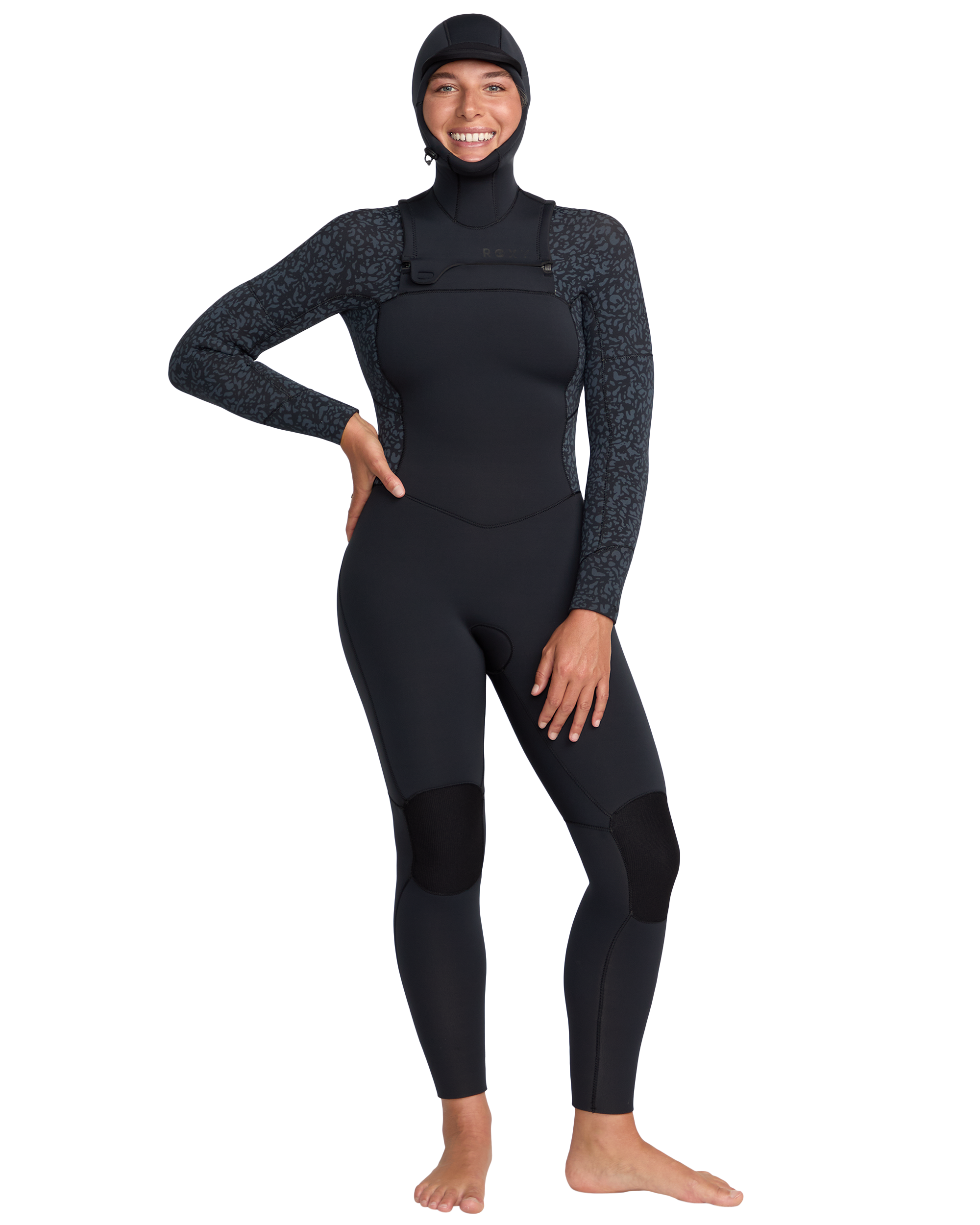 A person stands barefoot, smiling, and wearing the Roxy 5/4mm Swell Natural Hooded Front Zip Wetsuit with subtle patterned sleeves on a plain white background.