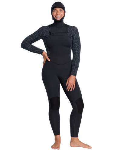 A person stands barefoot, smiling, and wearing the Roxy 5/4mm Swell Natural Hooded Front Zip Wetsuit with subtle patterned sleeves on a plain white background.