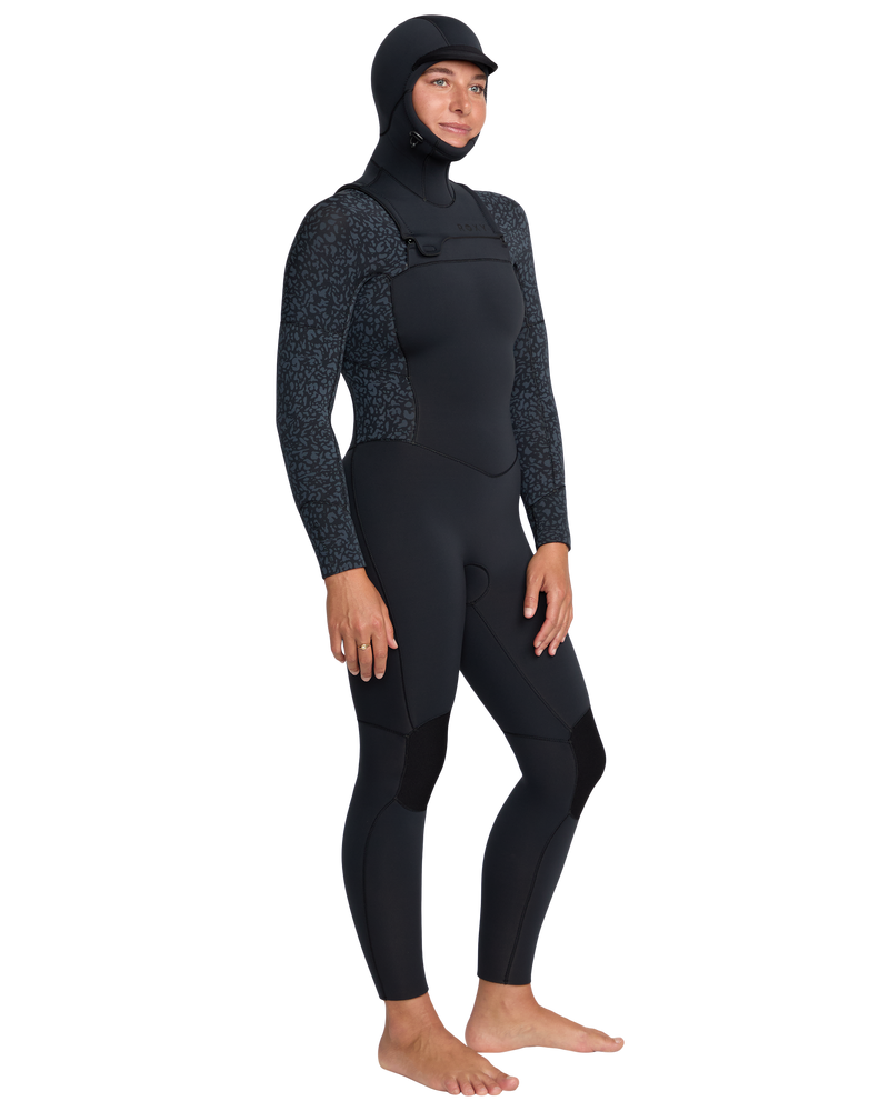 A person stands barefoot against a plain white background, wearing the Roxy 5/4mm Swell Natural Hooded Front Zip Wetsuit with patterned sleeves and an attached hood.