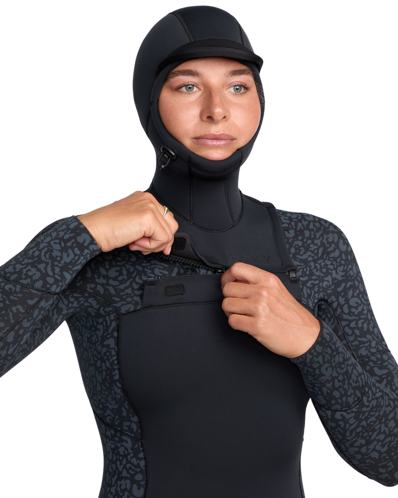 A person wearing the Roxy 5/4mm Swell Natural Hooded Front Zip Wetsuit in black with a subtle sleeve and shoulder texture pulls up the front zipper. This neoprene-free, natural rubber wetsuit has an attached hood. The background is plain white.