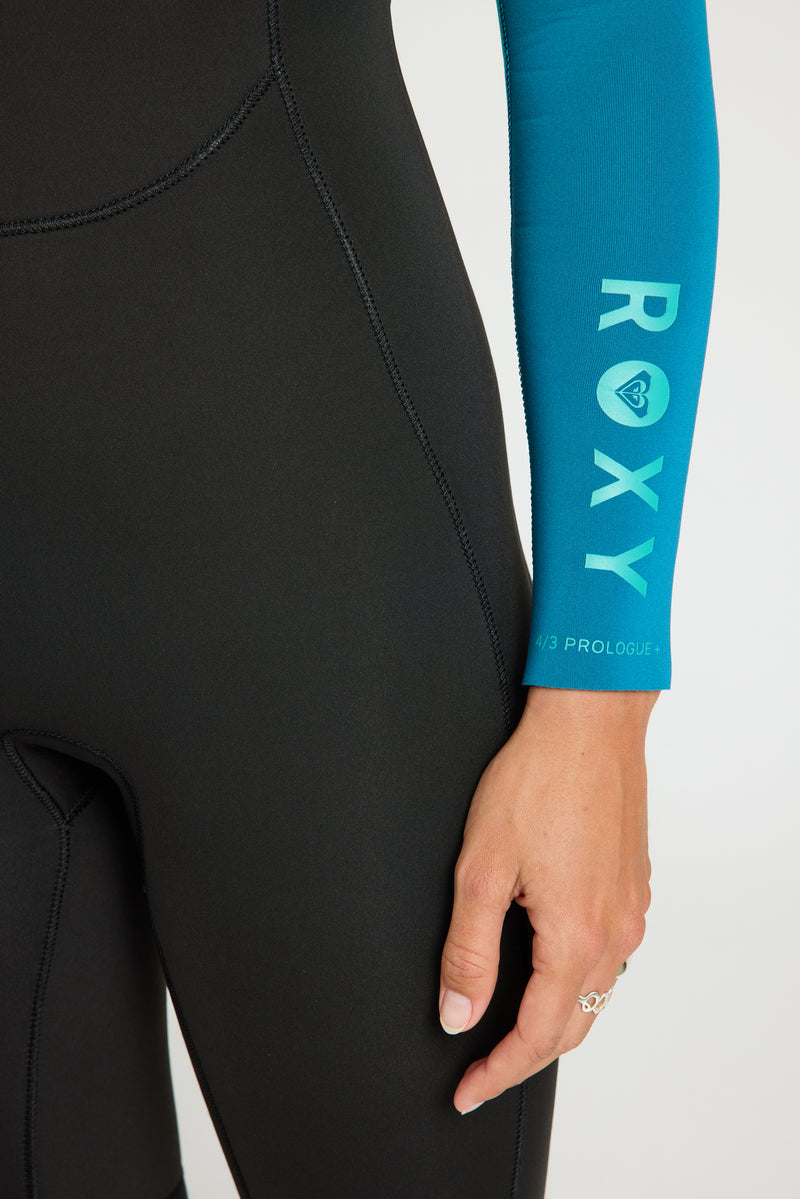 A close-up of a person wearing the Roxy 4/3mm Prologue GBS Back Zip Wetsuit, featuring a black body and a blue sleeve with "ROXY" printed on it. Their hand, adorned with a ring, rests near their hip.