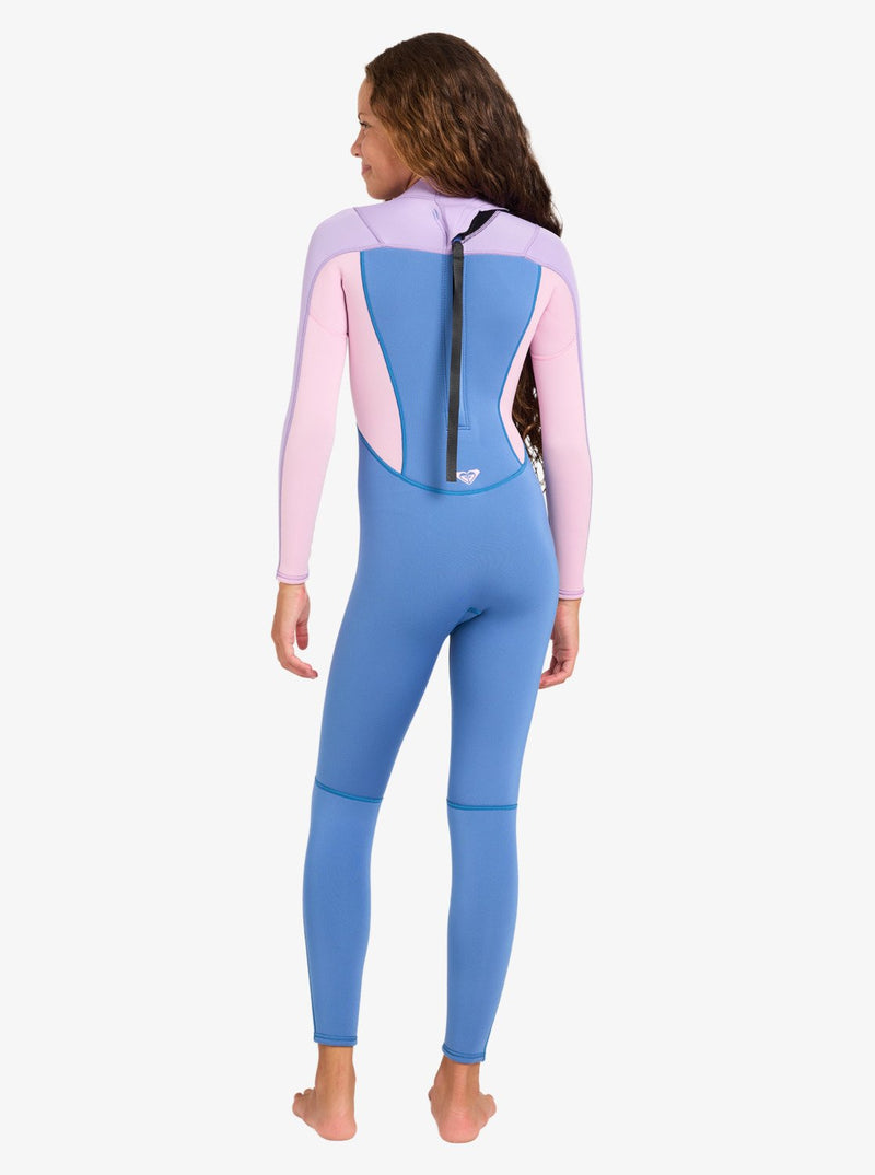 A young girl with long wavy hair stands barefoot and faces away, modeling the Roxy 4/3mm Prologue Girls’ Back Zip Wetsuit. The wetsuit features a blue and pink color-blocked design with a visible back zipper, shown against a plain white background.