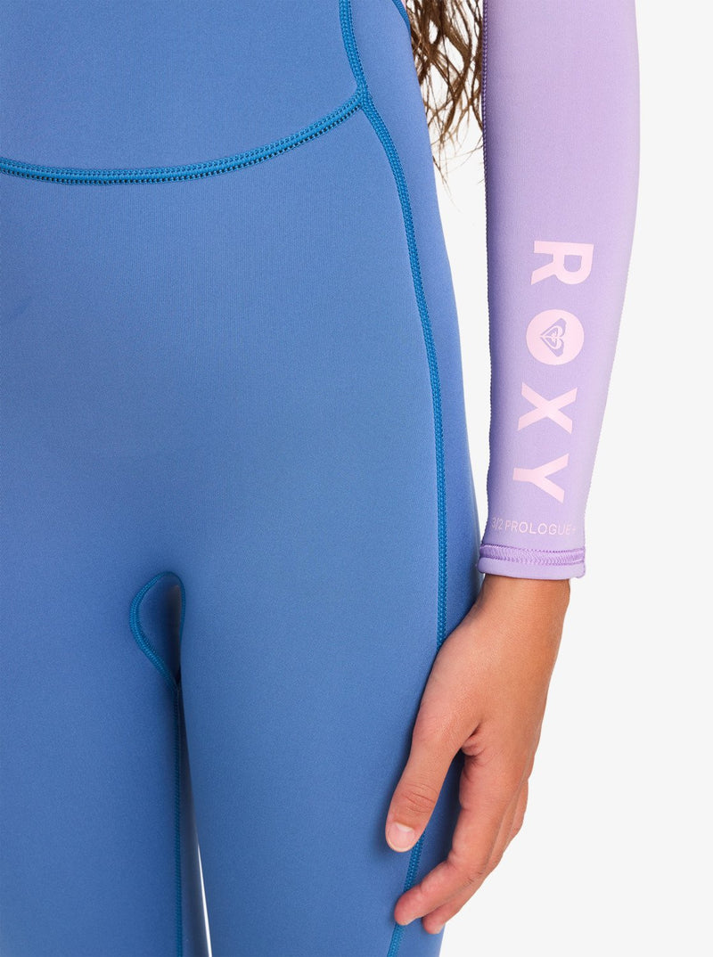 Close-up of a person wearing the Roxy 4/3mm Prologue Girls’ Back Zip Wetsuit in blue, featuring a lavender sleeve with “ROXY” and the brand’s logo printed in white. Only part of the person’s torso, right arm, and hand are visible.