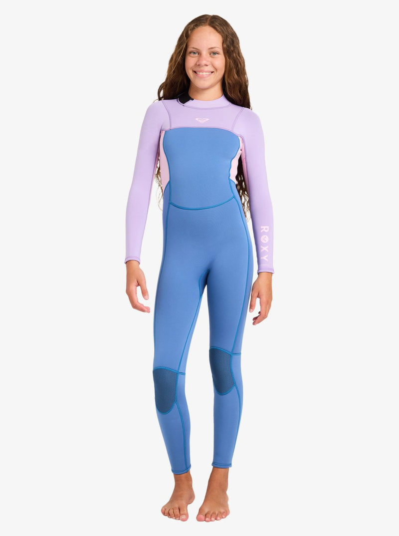 A girl stands barefoot and smiling on a white background, wearing the Roxy 4/3mm Prologue Girls’ Back Zip Wetsuit, which features a long-sleeved design with blue legs and torso, and light purple sleeves.