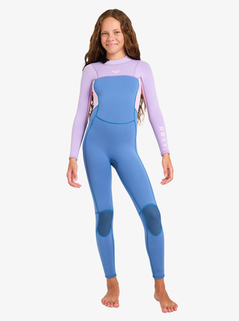 A young girl with long brown hair is seen barefoot and smiling against a plain white background, wearing the Roxy 4/3mm Prologue Girls’ Back Zip Wetsuit in shades of blue and lavender.