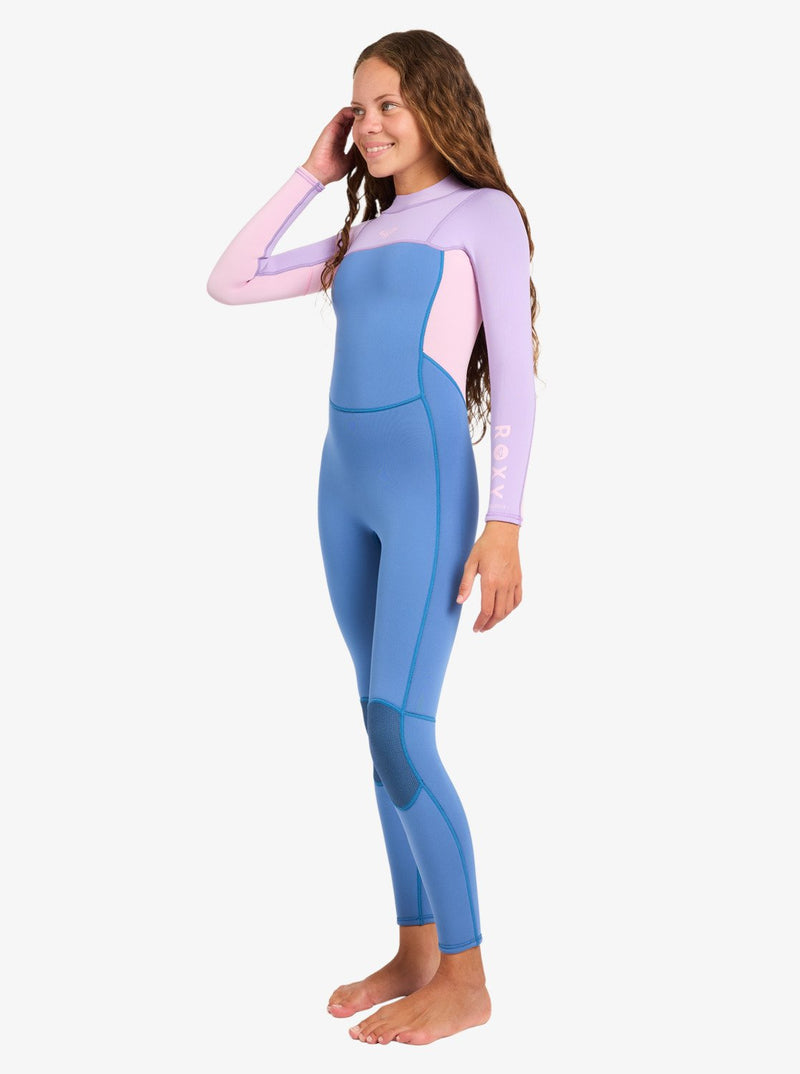 A woman with long wavy hair stands barefoot and smiles with one hand touching her hair, modeling the Roxy 4/3mm Prologue Girls’ Back Zip Wetsuit in blue and light purple against a plain white background.