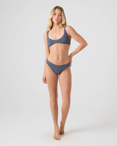 A woman with long blonde hair stands barefoot against a light background, confidently modeling the Roxy Haleiwa Hipster Full Bikini Bottom in textured blue made from recycled fibers.