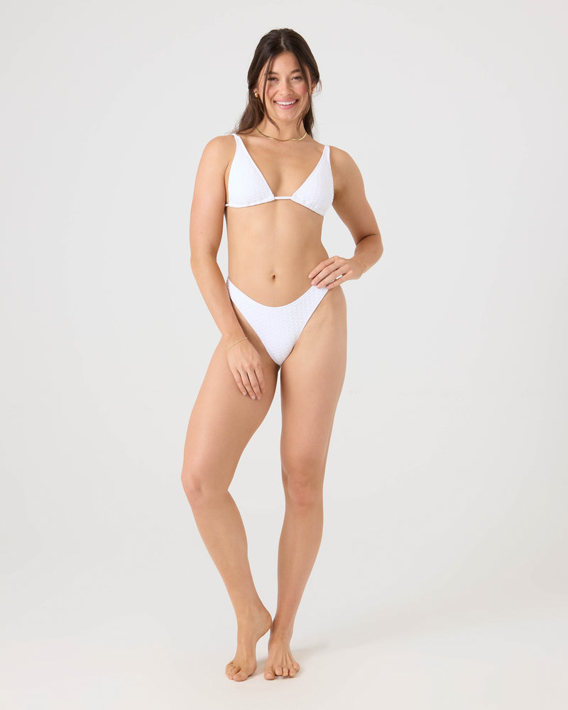 A woman with long brown hair stands barefoot, smiling, and wearing the Roxy Haleiwa Scoop Cheeky Bikini Bottom in white against a plain light gray background.