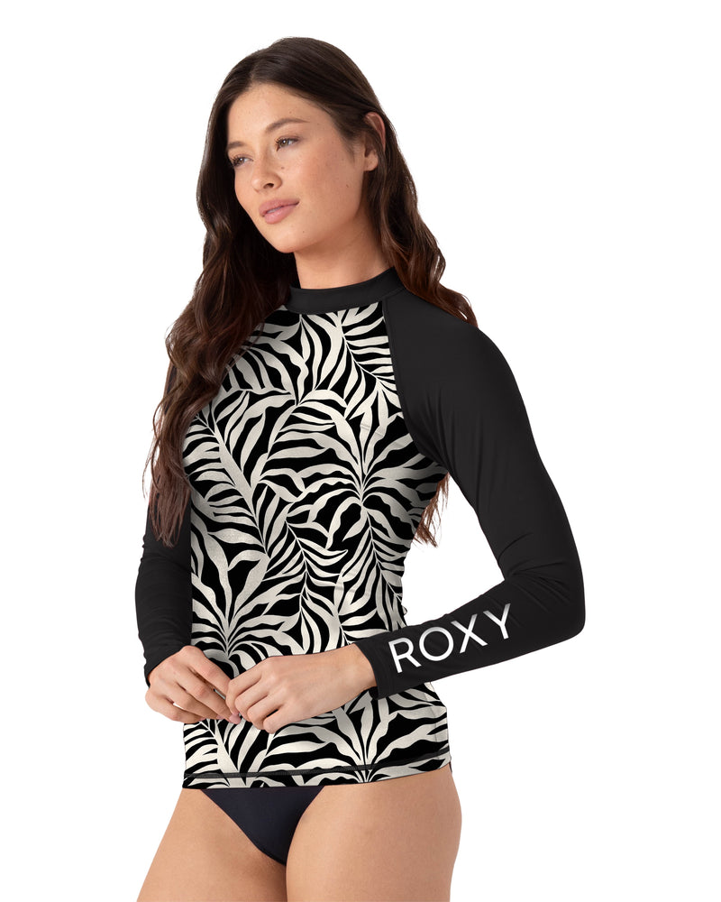 A woman models the Roxy Surfer's Paradise Rashguard, featuring black and white zebra print with "ROXY" on the sleeve, paired with black bikini bottoms. She stands to the side, her long brown hair flowing.
