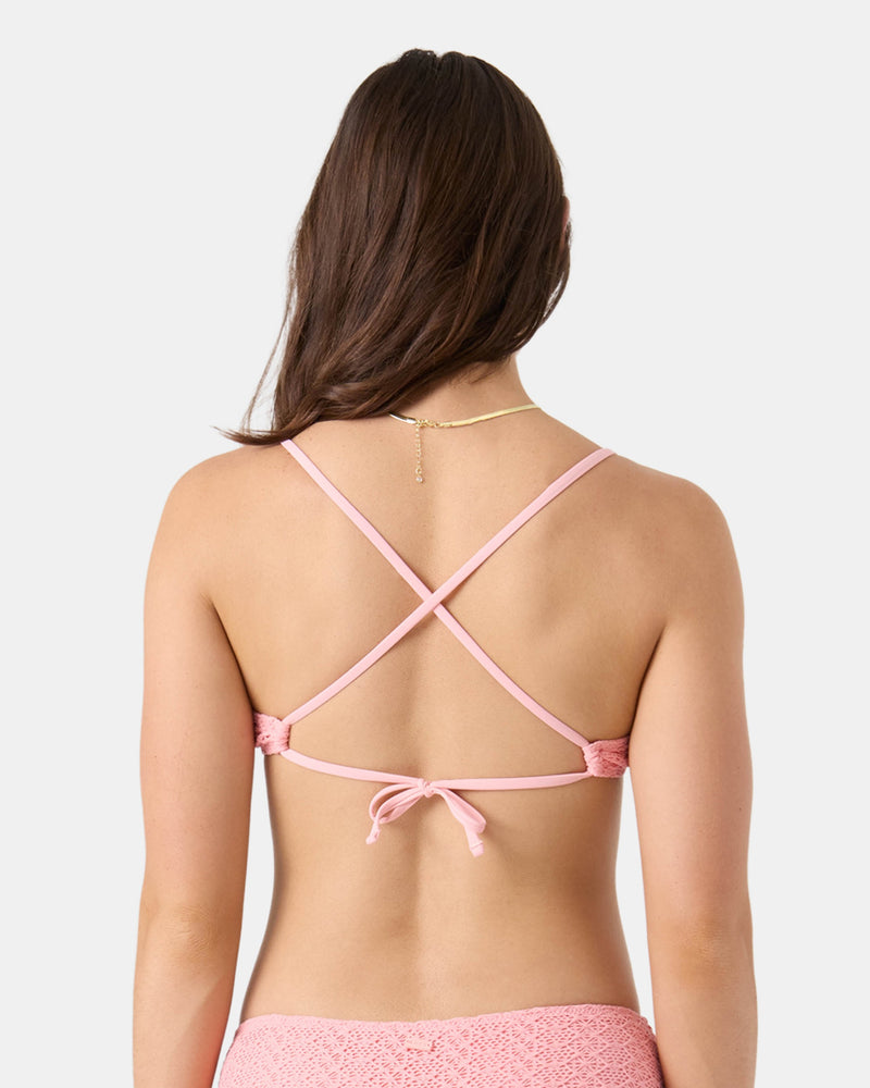 A woman with long brown hair models the Roxy Crochet Athletic Triangle Bikini Top in pink, featuring adjustable crisscross straps tied at the lower back. She wears a gold necklace and poses against a plain white background.