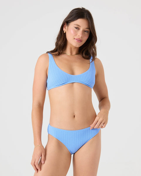 A woman with long brown hair wears the Roxy Haleiwa Scoop Bralette Bikini Top in textured light blue, made from recycled polyester with adjustable straps, paired with subtle jewelry, standing against a plain light background and smiling slightly.