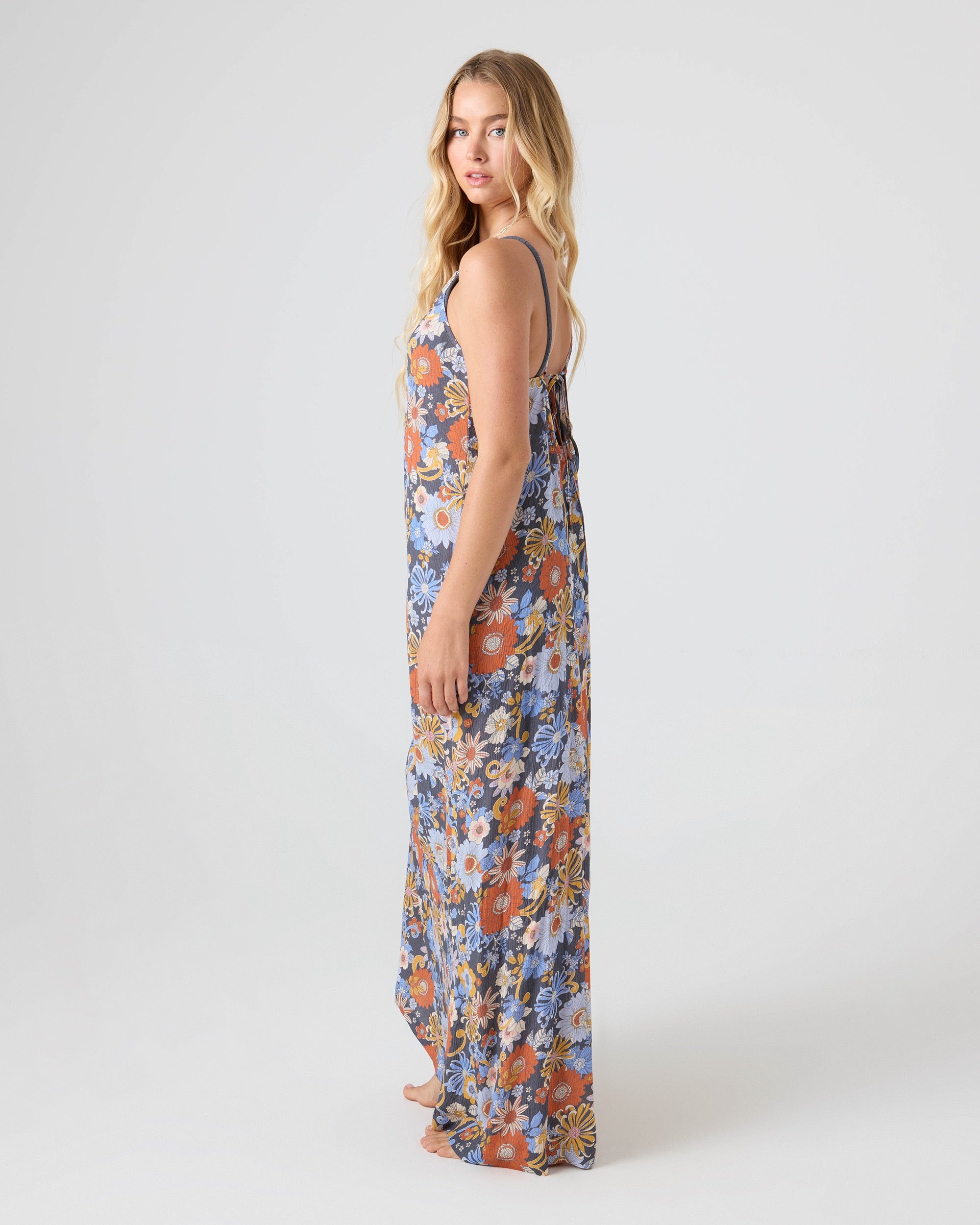 A woman with long blonde hair stands barefoot and sideways against a plain light backdrop, wearing the sleeveless, floor-length Soul Flower Cover Dress by Roxy.
