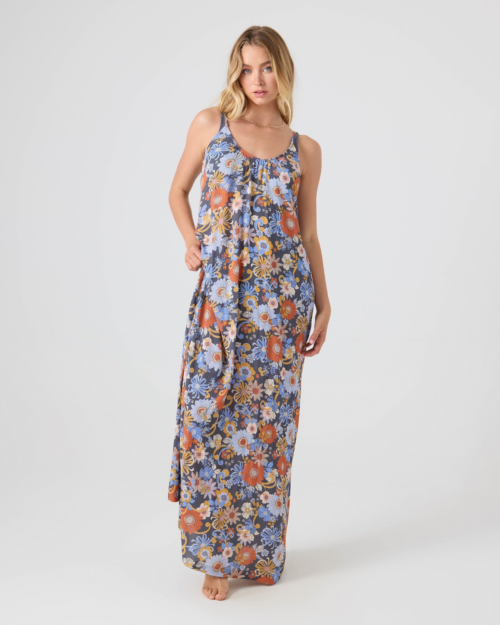 A blonde woman stands barefoot against a plain background in the Roxy Soul Flower Cover Dress—a blue, orange, and white floral maxi—holding her dress. This relaxed style is perfect as a beach cover-up.