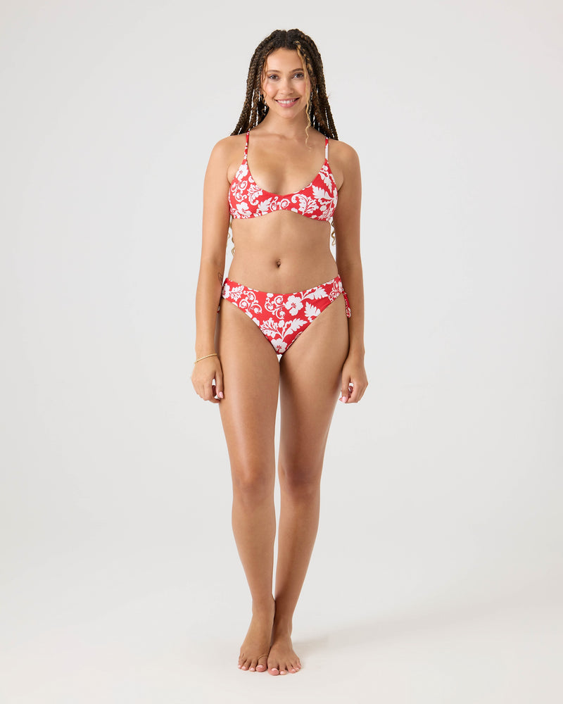 A woman with long braided hair stands barefoot against a plain background, modeling the Roxy Days Of Summer Athletic Triangle Bikini Top in Ivory Red and white floral print, smiling gently with her arms relaxed by her sides.