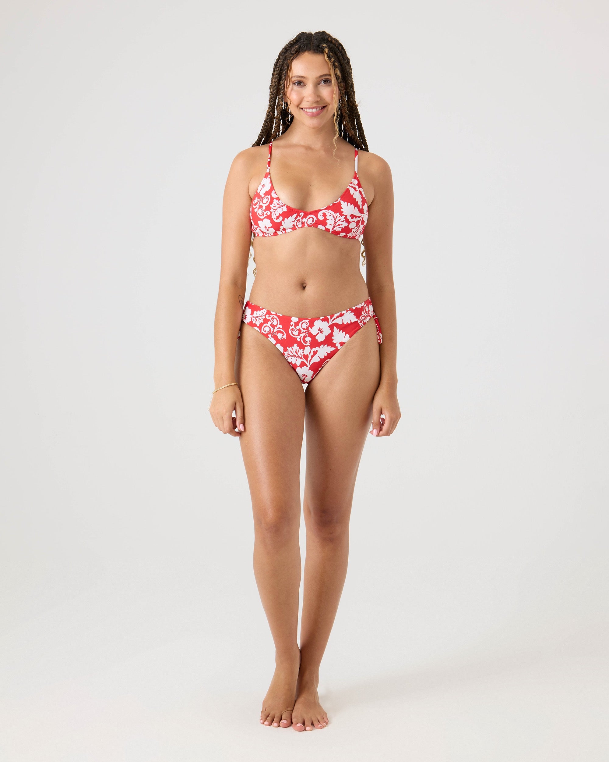 A woman with braided hair smiles while standing barefoot against a plain background, wearing the Roxy Days Of Summer Hipster Tie Side Bikini Bottom in a red and white floral print with a matching top.