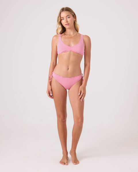 A woman with long blonde hair poses barefoot against a plain background, wearing the Roxy Good Vibes Hipster Lace Full Bikini Bottom in light pink and a matching scoop-neck top. She exudes a relaxed, natural vibe.