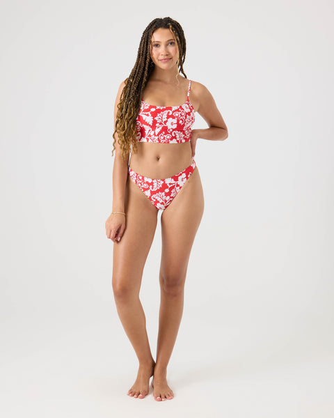 A woman with long braided hair poses barefoot against a plain background, smiling gently in the Roxy Days Of Summer Hipster Moderate Bikini Bottom, a red and white floral style made from recycled stretch fabric for comfortable, moderate coverage.