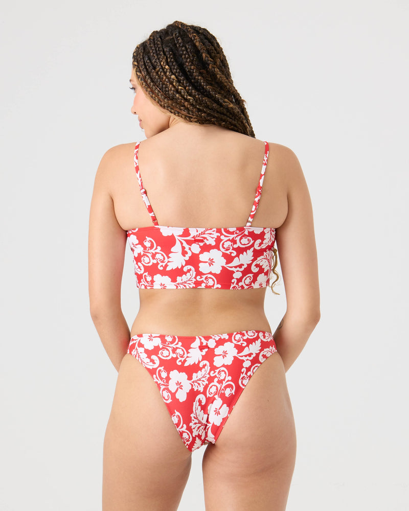 A woman with long, braided hair is seen from the back in a Roxy Days Of Summer Hipster Moderate Bikini Bottom, a high-waisted red and white floral style made from recycled stretch fabric. The background is plain and light-colored.