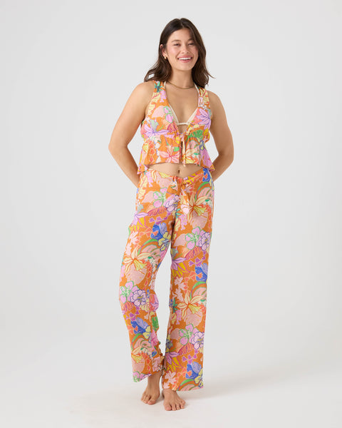 A woman stands barefoot against a plain background, smiling in Roxy's Lei It On Me Cover Up Pant—a high-waisted, wide-leg style in a vibrant Brown Multi floral print with orange, pink, purple, and green shades—perfect for stylish beachwear.