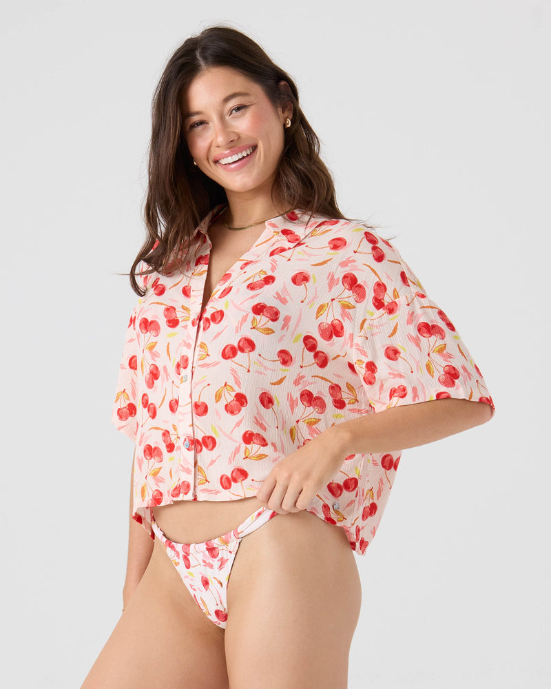 A woman smiles in the Roxy Cherry On Top Crop Button Up Shirt and cherry print bikini bottoms, standing against a plain light backdrop.