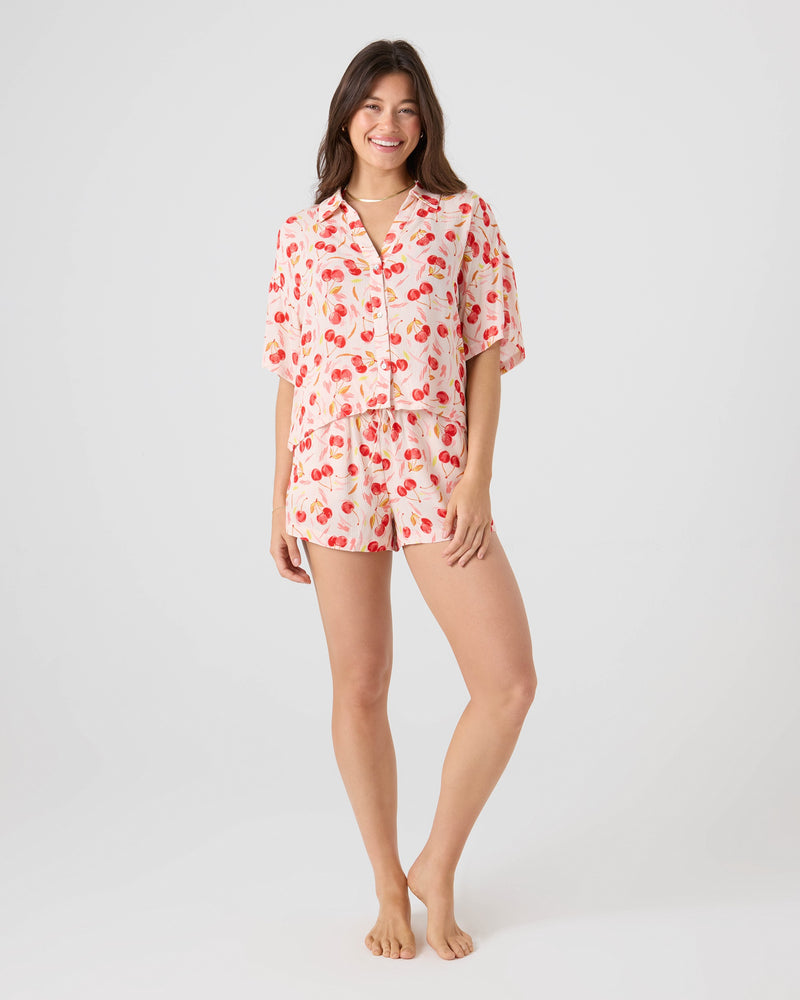 A woman stands smiling, wearing the Roxy Cherry On Top Crop Button Up Shirt with a vibrant cherry print and shorts pajama set, against a plain light background.