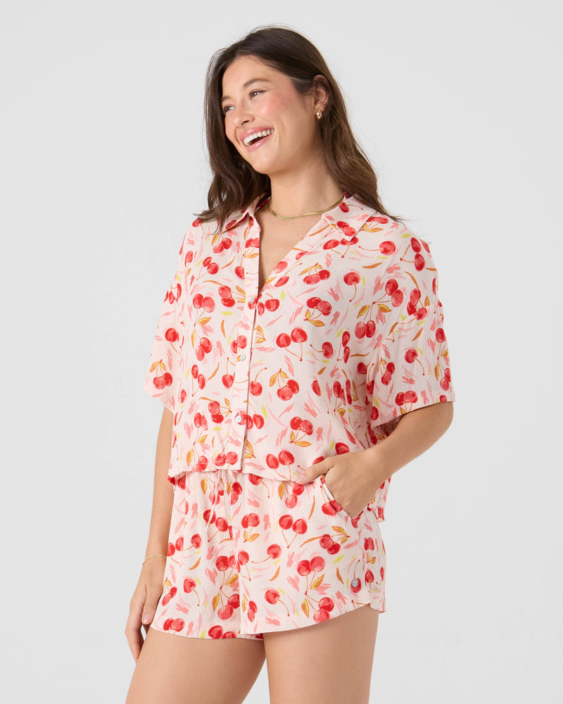 A woman smiles in the Roxy Cherry On Top Crop Button Up Shirt, featuring a vibrant red color with pink and red cherry print, paired with matching shorts. She stands confidently against a plain light background with one hand in her pocket.