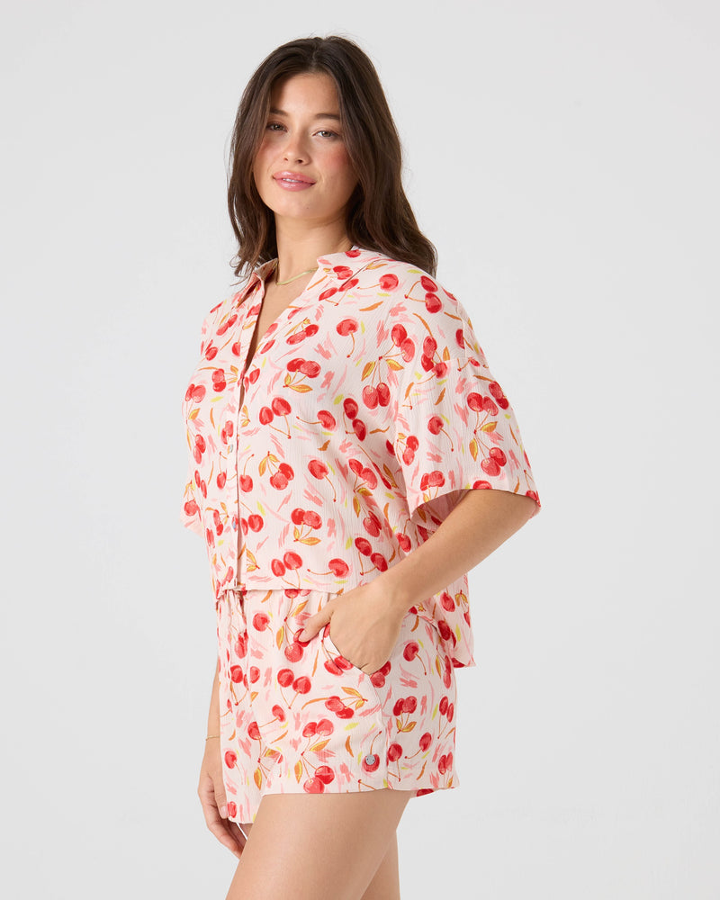 A woman wears the Roxy Cherry On Top Crop Button Up Shirt, a light pink top with a bold red cherry print, paired with matching shorts. She poses with one hand in her pocket against a plain background.