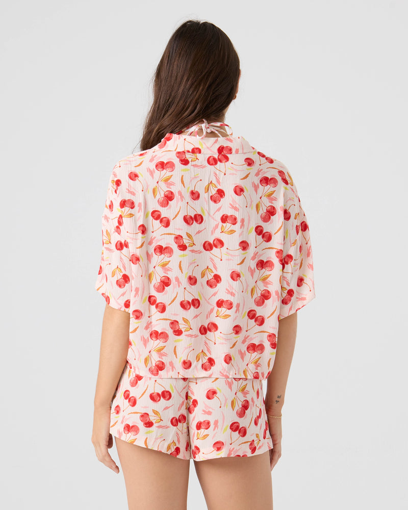 A woman with long brown hair is seen from behind wearing the Roxy Cherry On Top Crop Button Up Shirt and matching shorts pajama set, featuring a vibrant cherry print, against a light pink background.