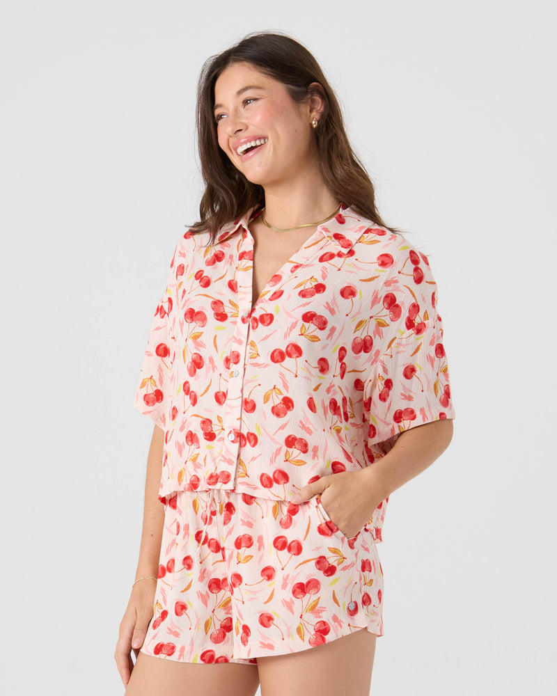 A woman smiles wearing the Roxy Cherry On Top Crop Stoke Short set, featuring a light pink, red floral short-sleeve button-up shirt and matching beach cover-up shorts. She stands with one hand in her pocket against a plain background.