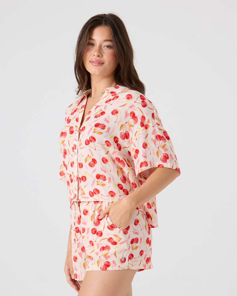 A woman with long brown hair stands sideways against a light background, wearing the Roxy Cherry On Top Crop Stoke Short and matching shirt in red cherry print, her hand in her pocket for an effortlessly stylish beach look.