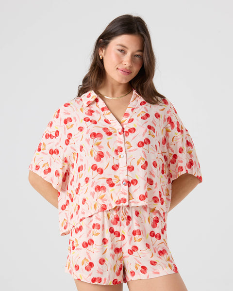A woman with long dark hair and an after-surf glow stands with hands on hips, wearing the Roxy Cherry On Top Crop Stoke Short set featuring a matching short-sleeve button-up shirt and shorts with a red cherry print on a light background.